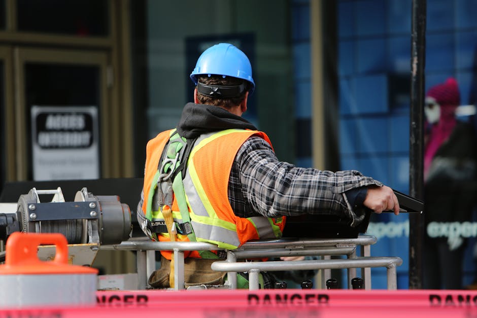 A construction worker wearing a blue helmet and safety vest operates equipment at an urban construction site